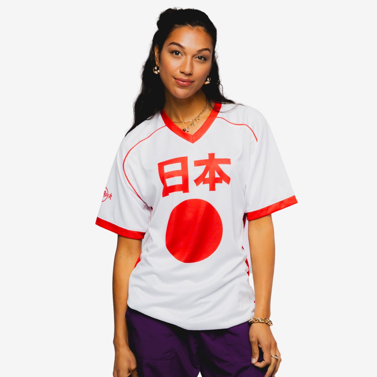 A young woman stands against a plain white background wearing a white sports jersey with red accents. Bold red Japanese characters and a large red circle are printed on the front of the shirt. She is also dressed in dark pants and accessorized with jewelry, including bracelets and earrings. The overall look suggests casual, sporty streetwear with a minimalist studio setting.