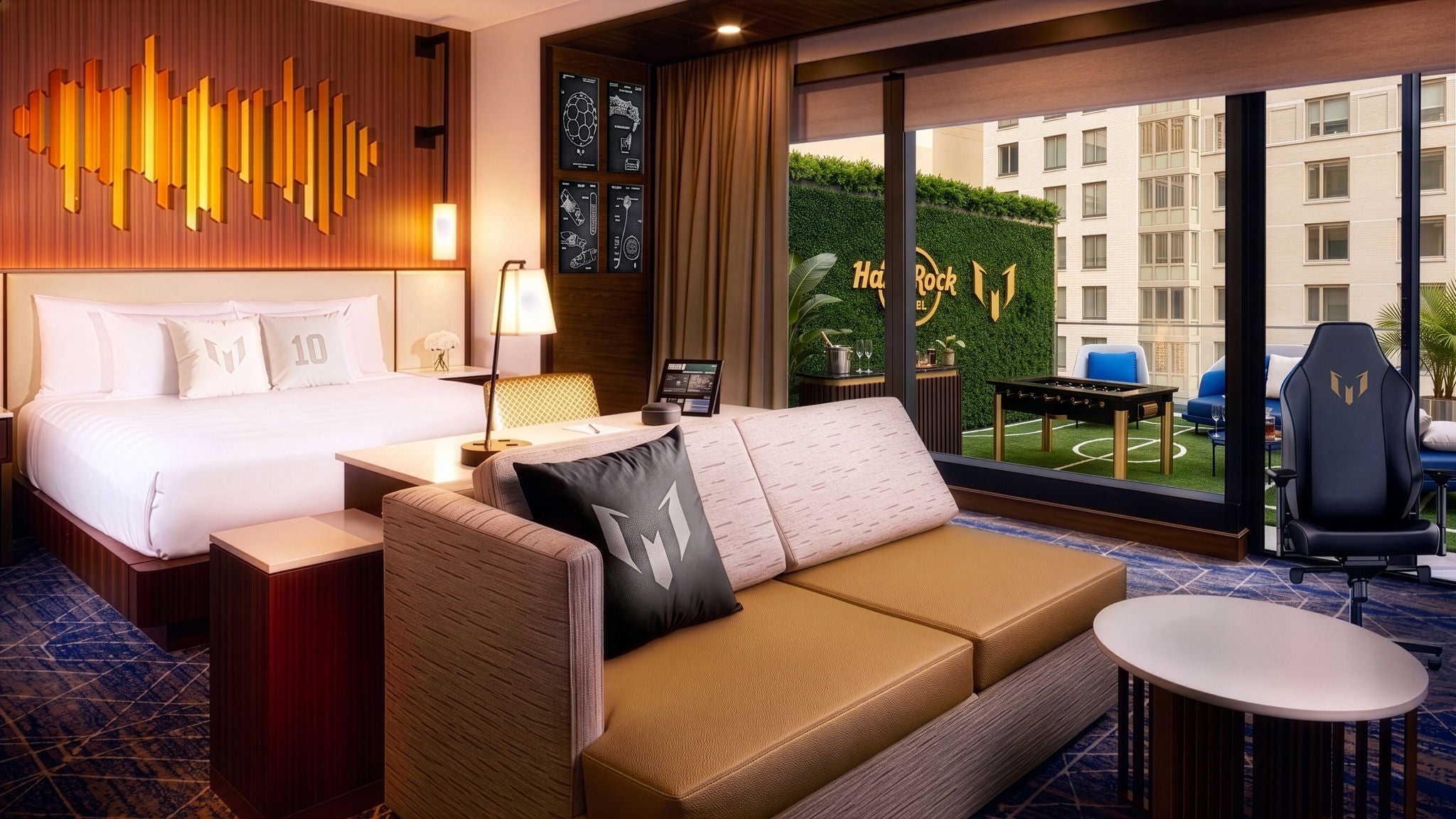 A contemporary hotel suite features a large bed, sofa, and work area in a warm, wood-accented interior. The room opens onto a terrace with green turf, outdoor seating, and a view of nearby buildings. A distinctive gaming-style chair and decorative pillows with a stylized "W" add a tech-forward, entertainment-focused feel. The words "Hard Rock" and the number "10" are visible on the wall art and pillows, reinforcing the themed design.