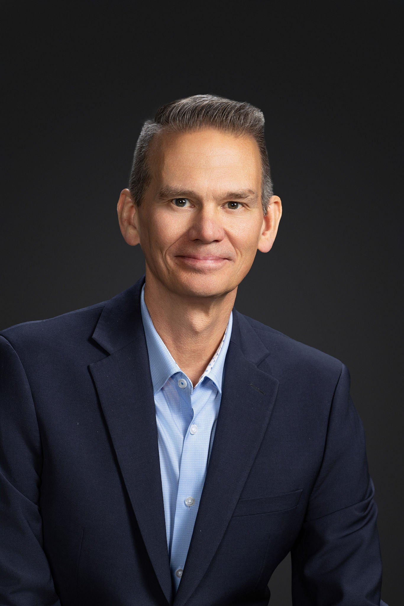 Portrait of a middle-aged man wearing a navy blue blazer and light blue dress shirt. He is seated and facing the camera with a slight smile, against a plain dark studio background. The image has a clean, professional look suitable for corporate or busines