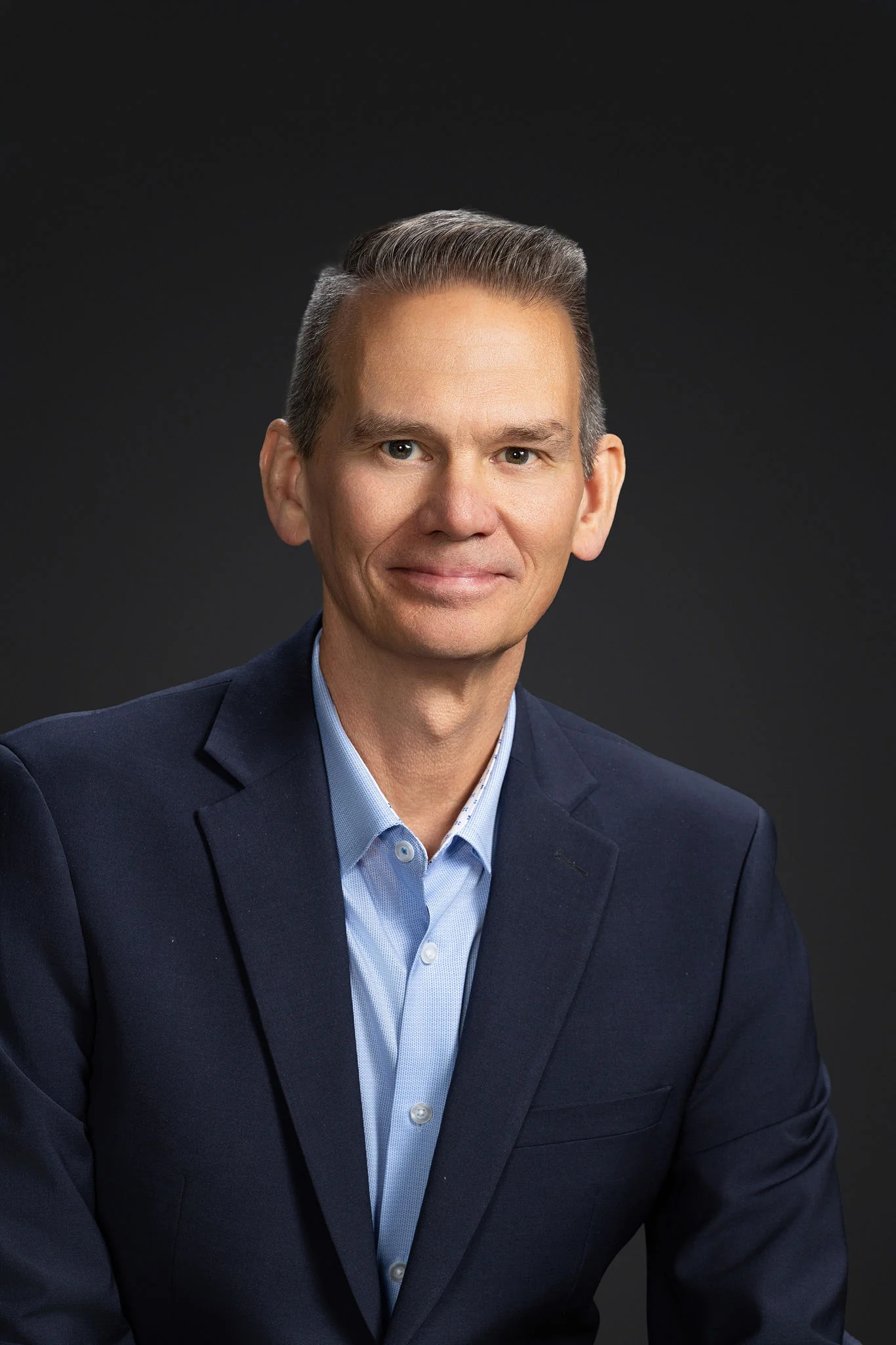 Portrait of a middle-aged man wearing a navy blue blazer and light blue dress shirt. He is seated and facing the camera with a slight smile, against a plain dark studio background. The image has a clean, professional look suitable for corporate or busines