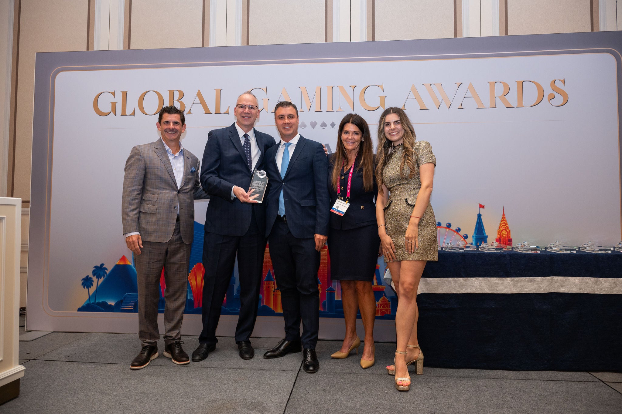 Five adults stand together on a stage at the Global Gaming Awards event. One man in the center holds a trophy while the others smile beside him. The backdrop features the text "GLOBAL GAMING AWARDS" and colorful cityscape illustrations. The setting appear
