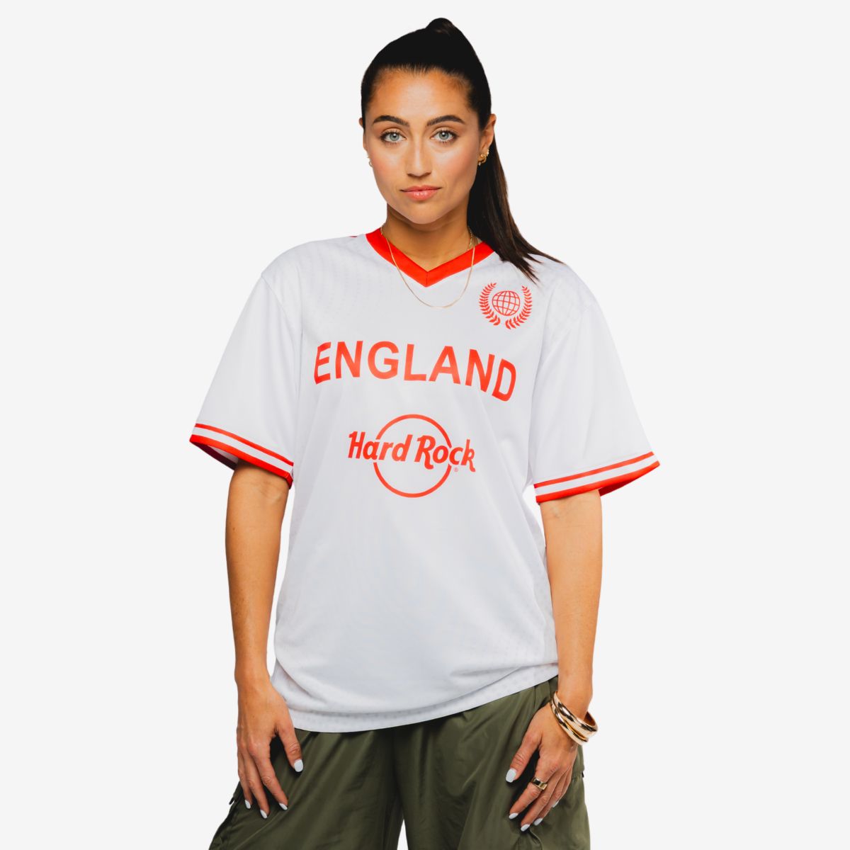 A person stands facing forward wearing a white England football-style jersey with red accents. The shirt prominently features the words "ENGLAND" and "Hard Rock" on the front, along with a circular emblem near the shoulder. The individual has long dark hair tied back and is dressed casually with loose green pants and bracelets. The background is plain and light, keeping focus on the jersey and its sporty, casual style.