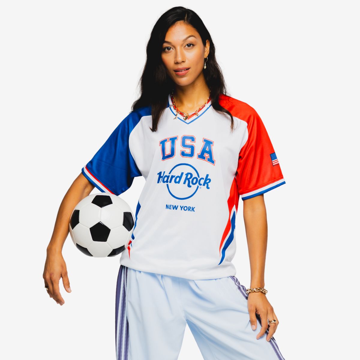 A young woman stands holding a classic black and white soccer ball at her side. She wears a white jersey with blue and red sleeves featuring the text "USA" and "Hard Rock" along with "New York" on the front. The outfit is sporty and casual, paired with light-colored pants against a clean white background. The overall scene emphasizes national pride and an energetic, athletic lifestyle.