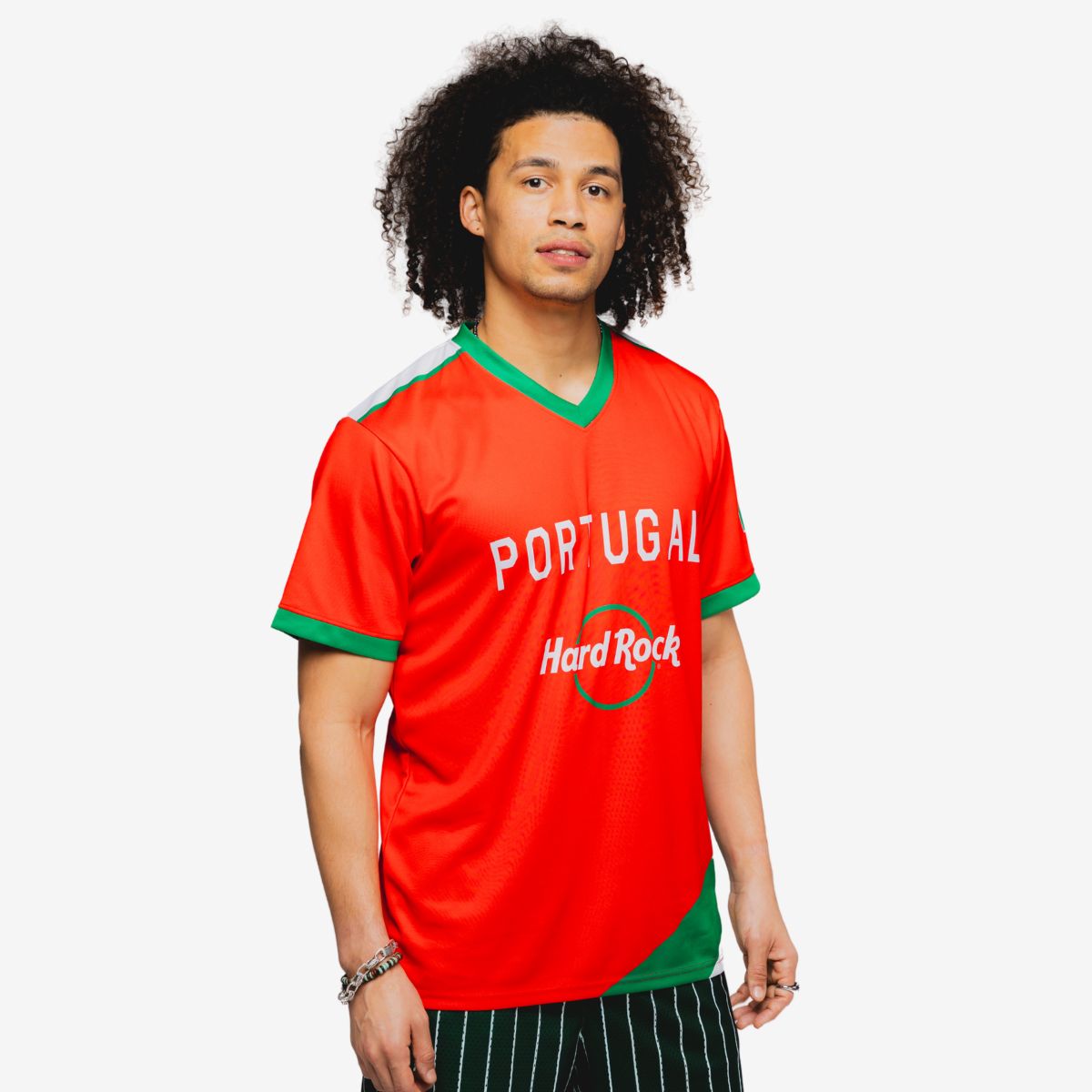 A person stands against a plain light background wearing a bright red Portugal Hard Rock soccer-style jersey. The shirt features green trim on the collar and sleeves with white text reading "PORTUGAL" and "Hard Rock" on the front. The subject has curly hair and is shown from about mid-thigh up, with their face blurred for privacy. The overall look is casual and sporty, with the vivid red and green colors standing out prominently.
