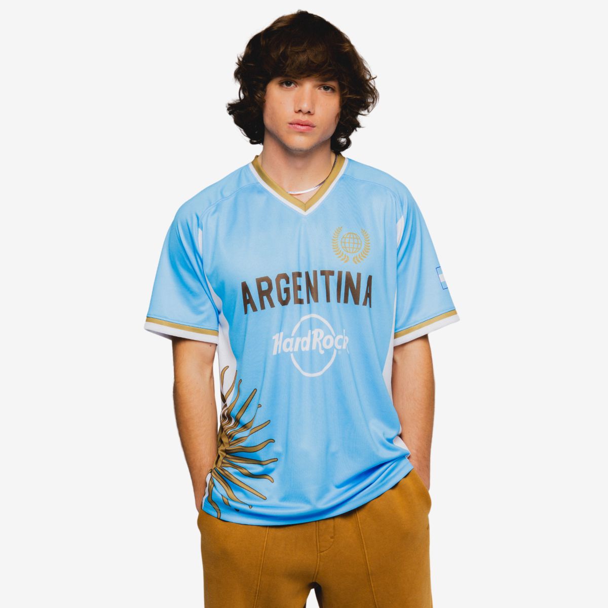 A person stands facing forward wearing a light blue Argentina soccer jersey. The shirt features the word "ARGENTINA" across the chest with additional decorative graphics including a stylized sun motif. The individual has medium-length hair and their face is blurred for anonymity. The background is plain white, drawing attention to the sportswear and its colors.